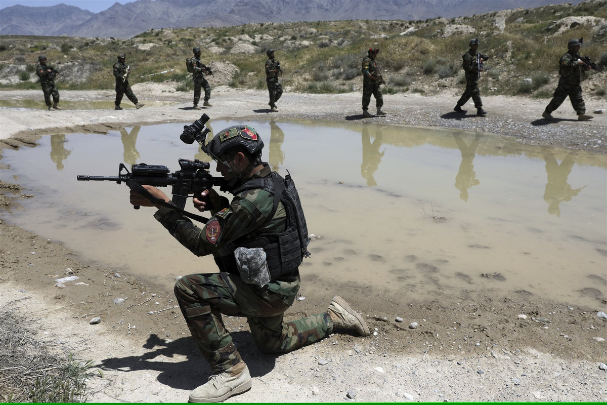 An Afghan National Army commando kneels with a rifle raised during a training exercise, with soldiers patrolling in the background.