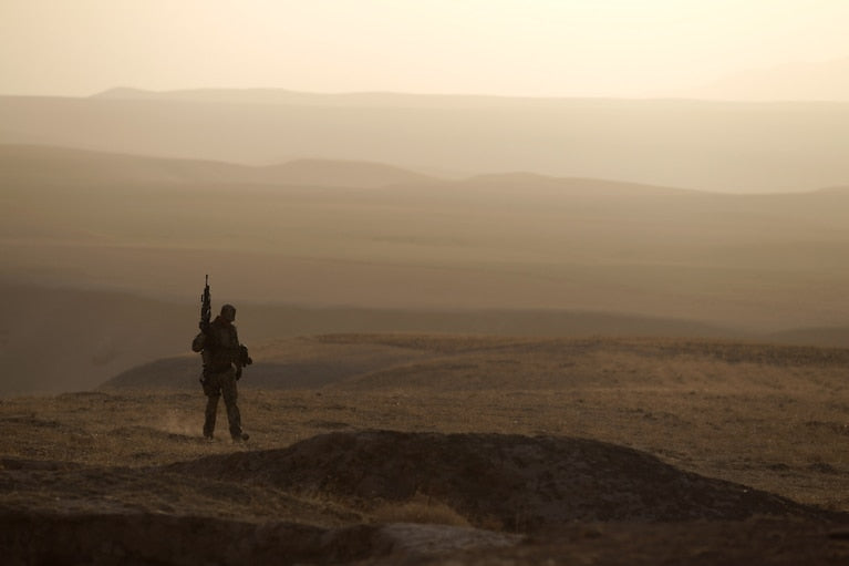 A lone soldier carrying a rifle stands silhouetted against a vast, hazy Afghan desert landscape at dusk.