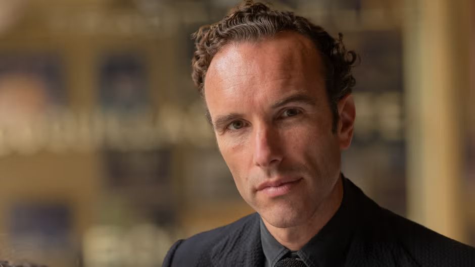 Elliot Ackerman portrait wearing a black suit and tie, photographed against a warm blurred background. Ackerman is a New York Times bestselling author, journalist, and former Marine Corps officer.